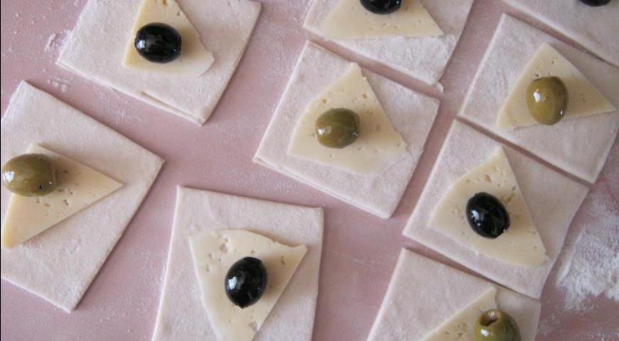 Тесто раскатываем и нарезаем на небольшие квадратики. На каждый кусочек выкладываем тонкие ломтики голландского сыра, а сверху — одну оливку или маслину. Сворачиваем тесто конвертиком, аккуратно защипывая края.