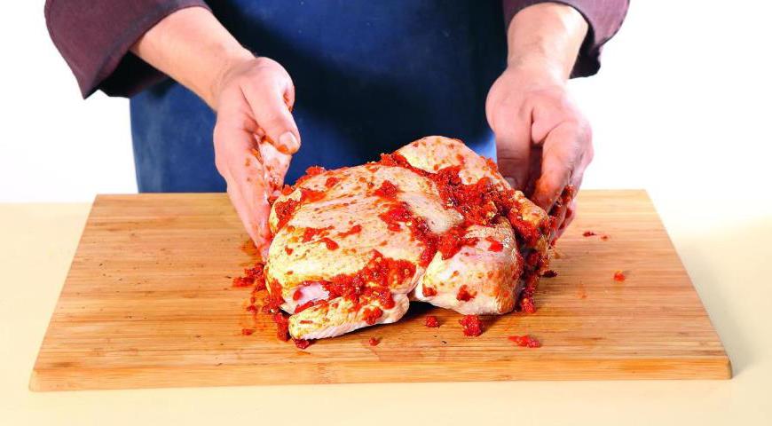 Очистите курицу от лишнего жира и железы на гузке, подверните крылышки для более аккуратного вида. Намажьте курицу аджикой внутри и закрепите ножки кулинарной нитью. После этого тщательно натрите птицу аджикой снаружи со всех сторон. Уложите курицу на овощи грудкой вниз.