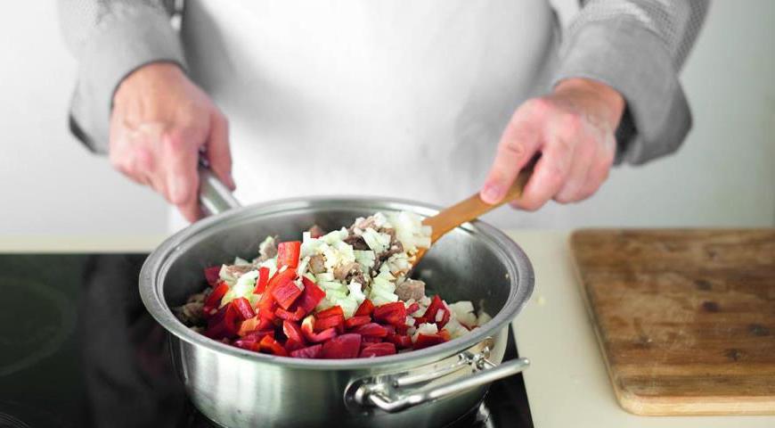 Верните все мясо в кастрюлю, добавьте лук, чеснок и сладкий перец. Перемешайте, добавьте острый красный перец и еще раз перемешайте. Накройте крышкой и тушите на среднем огне, периодически помешивая, 25 минут.