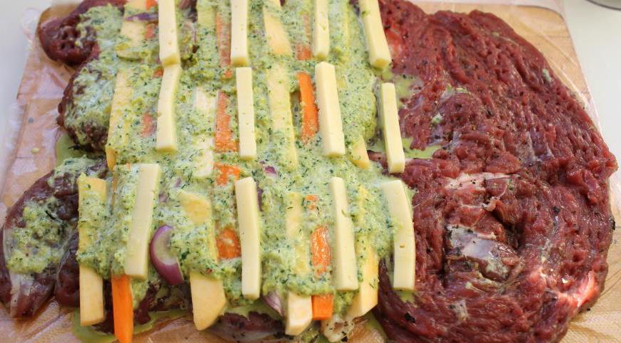 Разверните мясо, оставив его на плёнке. Выложите начинку: чередуйте репу, морковь, лук и сыр. Немного овощей оставьте для дальнейшего использования. Поверх начинки нанесите примерно две трети приготовленного соуса.