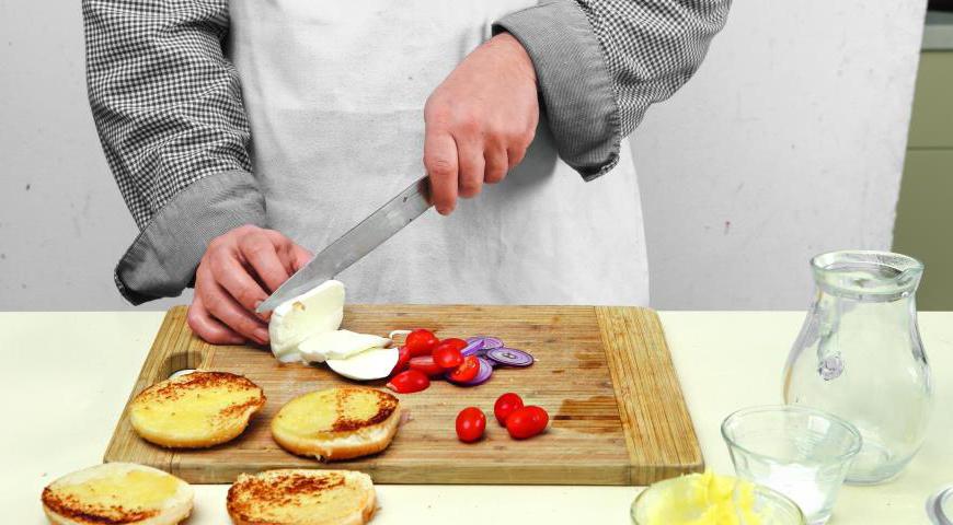 Смешайте готовый фарш с желтком, замороженным жиром и обработанным луком. Посолите и поперчите по вкусу. Сформируйте 4 котлеты, которые будут немного больше булочек. Выложите их на пергамент и отправьте в холодильник на 35 минут. За это время приготовьте булочки: разрежьте их пополам, смажьте маслом и обжарьте до золотистого цвета. Нарежьте томаты и моцареллу тонкими слоями.