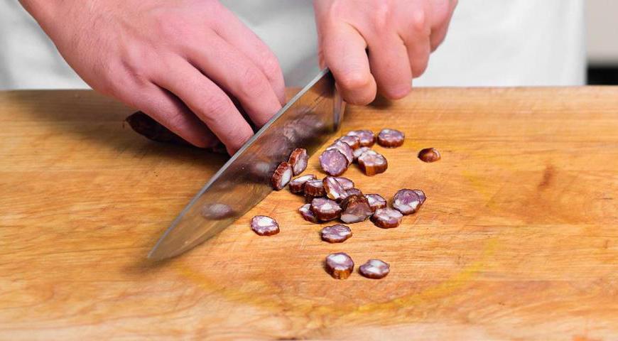 Нарежьте колбаски кружочками и положите их в глубокую кастрюлю или горшок. Добавьте промытую фасоль, обжаренные овощи и томатную массу. Залейте 220 мл горячей воды, посолите, поперчите и добавьте любимые специи.