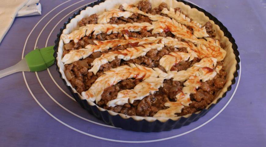 Выложить мясную начинку по центру. Из оставшегося теста вырезать полоски и уложить их сверху в виде решетки-ёлочки. Смазать верх томатным кетчупом для красивого цвета.