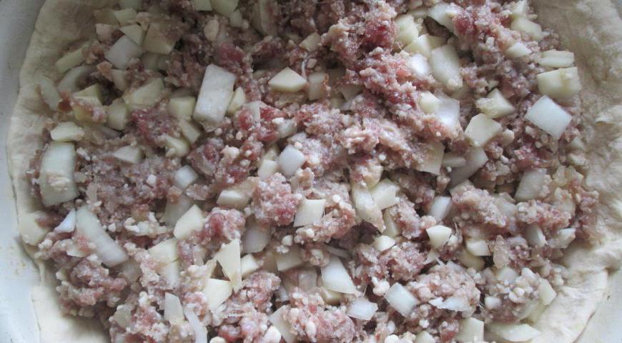 Пока тесто отдыхает, займемся начинкой. Картофель очистите и нарежьте мелкими кубиками (примерно 1–1,5 см), лук нашинкуйте мелко. Добавьте к ним говяжий фарш, посолите, поперчите по вкусу и хорошо перемешайте. За это время тесто должно немного «отдохнуть». Возьмите его и еще раз обомните. Разделите на три части в пропорции 1:3 — меньшая часть будет использоваться для кендека (маленькой заглушки) и крышки, большая — для основания пирога. Раскатайте большую часть теста так, чтобы её диаметр был больше формы (сковороды или противня). Выложите начинку в форму, затем аккуратно поднимите края теста и защипите их в центре, формируя «мешок».